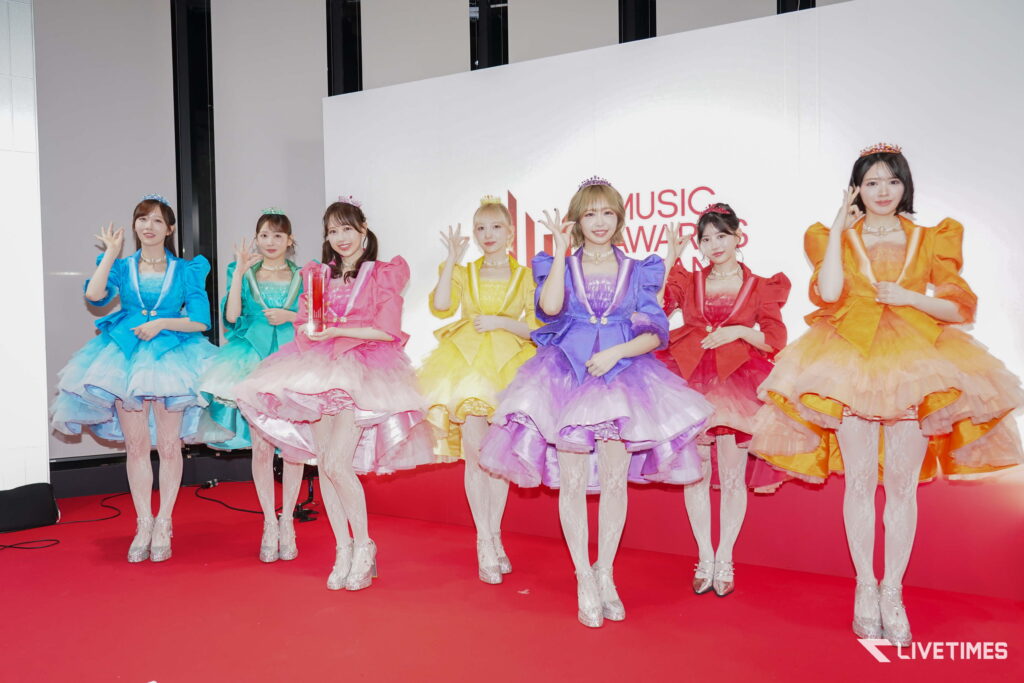 Seven women in bright, ruffled, princess-style dresses pose on a red carpet at a music awards event, waving at the camera and wearing tiaras.