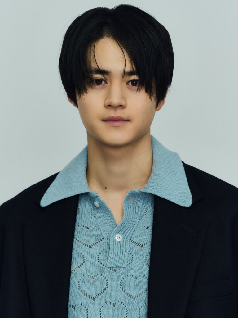 Head-and-shoulders portrait of a person with short black hair, wearing a light blue knit polo and dark blazer against a pale background