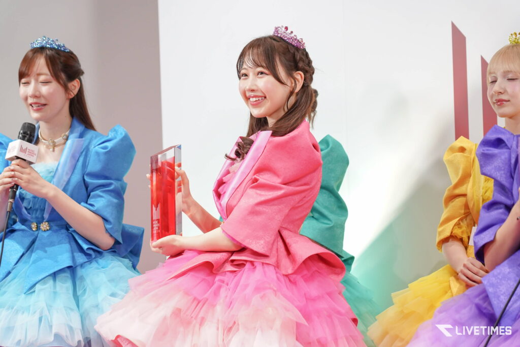 Three women in colorful gowns with tiaras on stage; the center woman in pink holds a glass award with a smile.