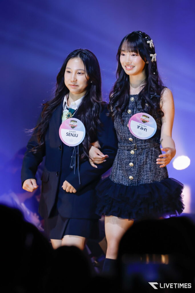 Two young women walk arm in arm on stage, wearing badges and smiles, under purple spotlight.