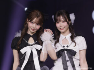 Two young women in matching black‑and‑white outfits pose on stage, holding hands and smiling.