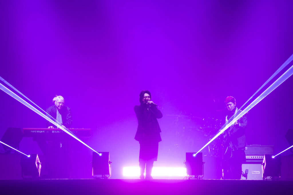 Band performing on a purple-lit stage with a singer center and keyboardist/guitarists on either side, laser beams crossing the scene.