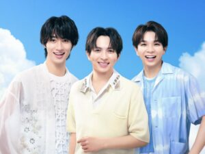 Three smiling men pose together outdoors against a bright blue sky, wearing light casual outfits.