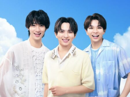 Three smiling men pose together outdoors against a bright blue sky, wearing light casual outfits.
