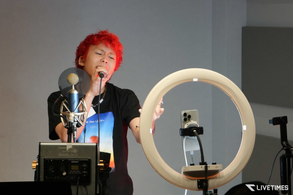 Singer with bright red hair sings into a microphone in a recording studio, with a pop filter nearby and a ring light visible.