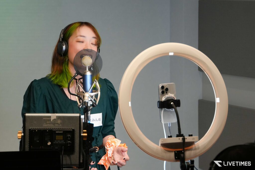 Woman in teal blouse sings into a mic with pop filter in a recording studio, wearing headphones; ring light and audio gear nearby.