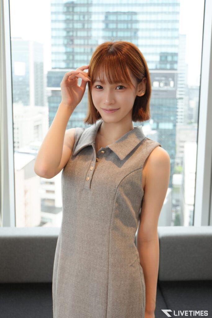 Young woman with short brown hair in a gray sleeveless dress poses indoors by a large window with a cityscape behind her, hand touching her hair.