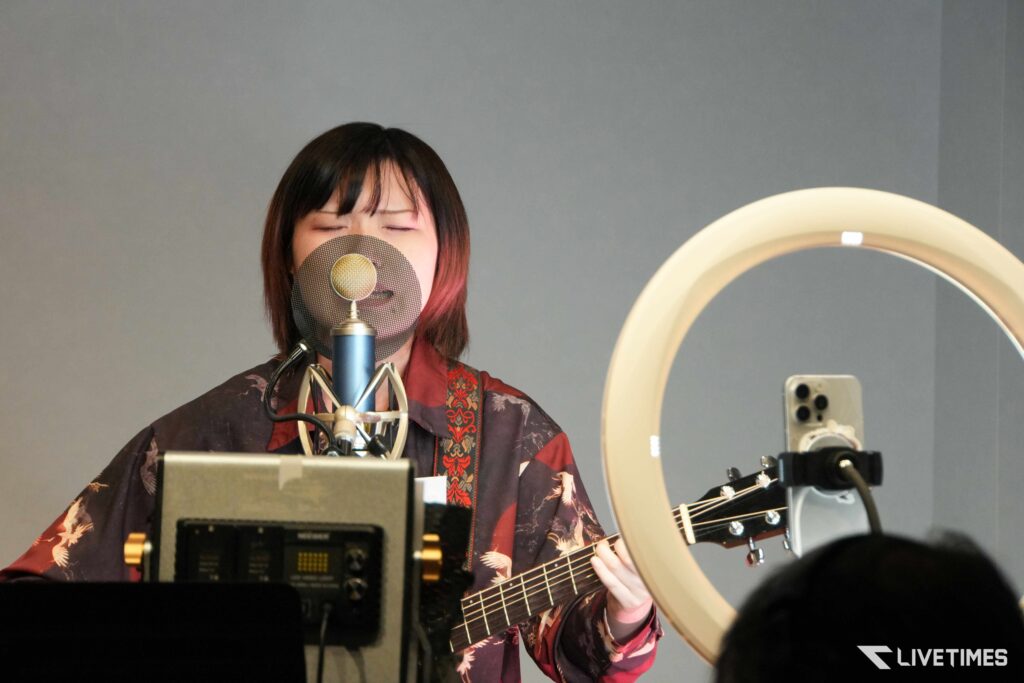Female singer wearing a red and black patterned kimono sings into a blue studio microphone with a circular pop filter in front of her face; ring light visible on the right.