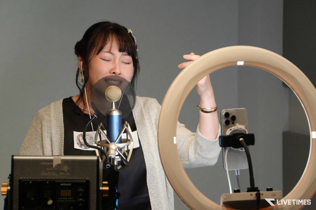 Woman singing into a studio microphone with a pop filter, wearing a light cardigan and headphones in a recording setup.