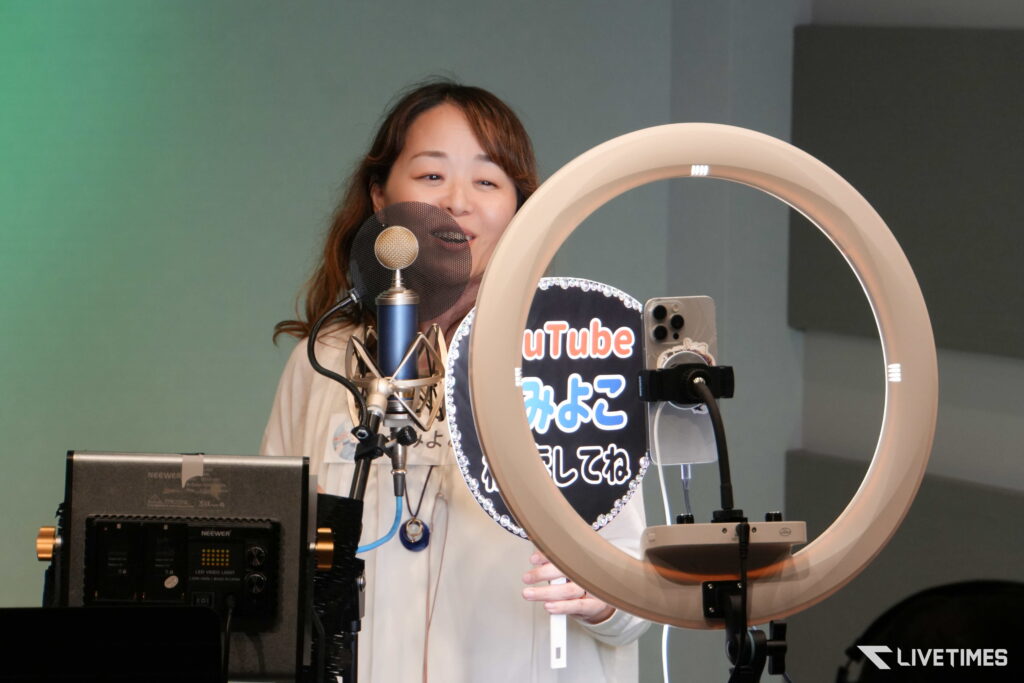 Woman in a recording studio speaks into a pop-filtered microphone, with a large ring light nearby.