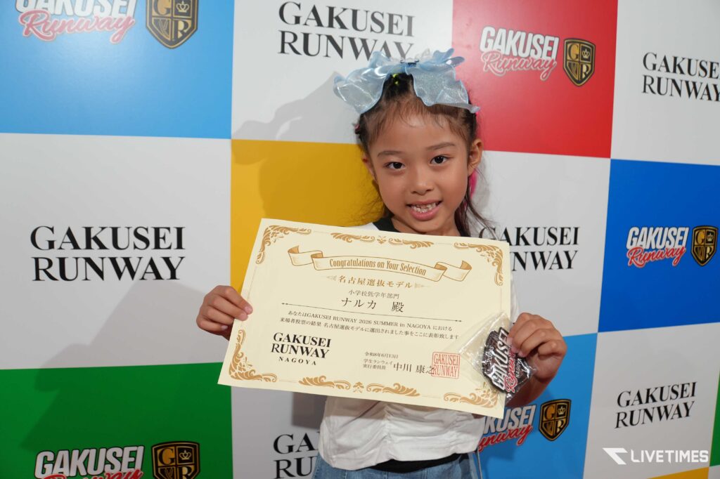 Young girl with a blue bow holds a large certificate at a GAKUSEI RUNWAY backdrop.