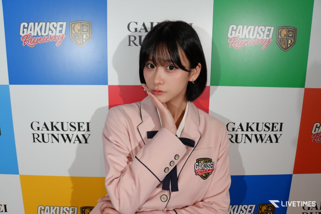 Young woman in a pale pink blazer posing with her hand under her chin, against a colorful GAKUSEI RUNWAY backdrop.