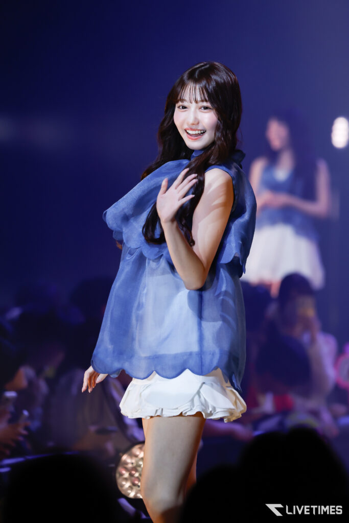 Smiling female performer on stage wearing a blue sleeveless top and white ruffled skirt, against a dark blue background.