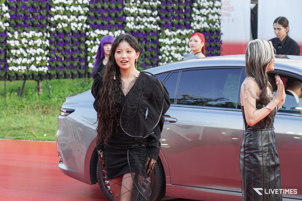 Woman in a black outfit stands near a silver car on a red carpet with a purple-white flower wall in the background, at an outdoor event.