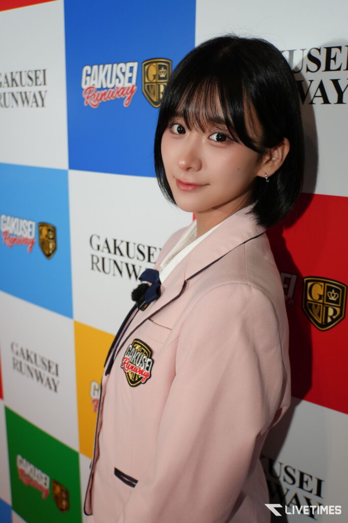 Young woman with short black hair wearing a pink blazer posing in front of a 'Gakusei Runway' event backdrop