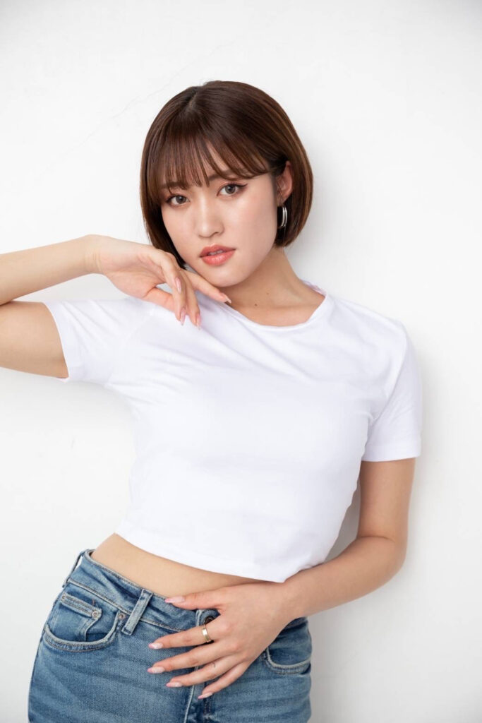Portrait of a young woman with a brown bob haircut, wearing a white t-shirt and blue jeans, posing with her hand near her chin against a white background.