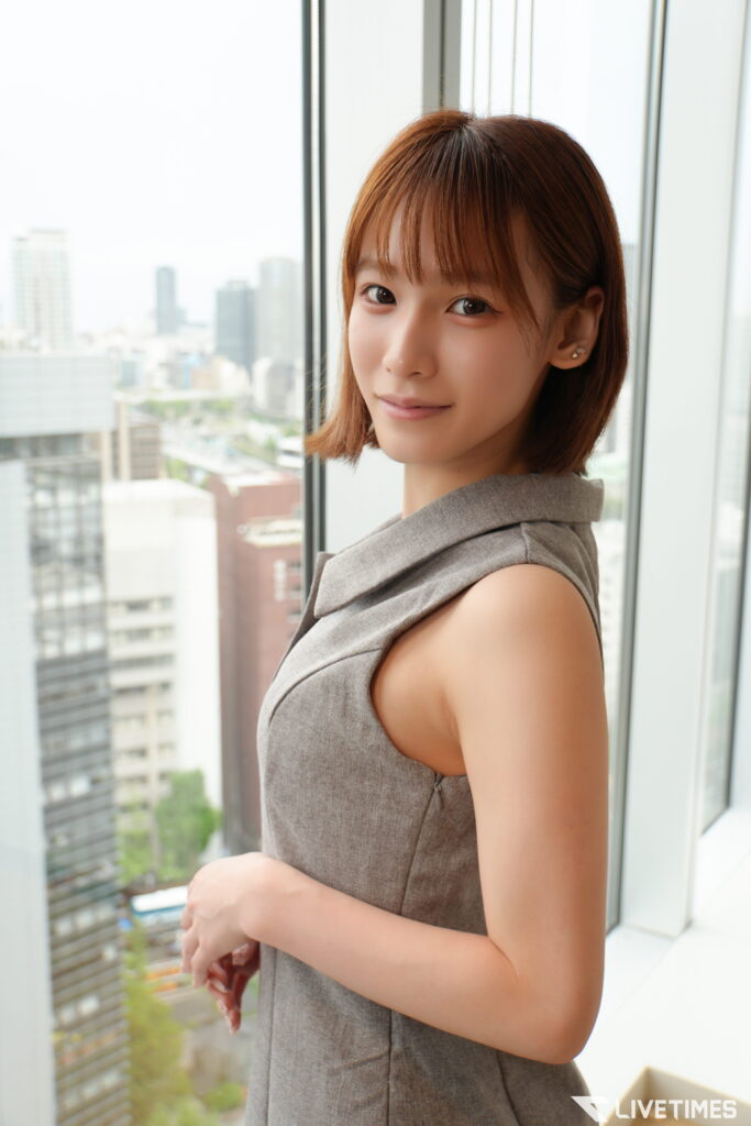 Portrait of a young woman with short brown hair wearing a gray sleeveless dress, standing by a window with a city skyline in the background, smiling slightly.
