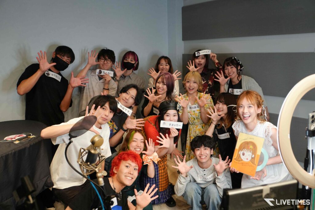 A large group of about twenty people in a studio, smiling and waving at the camera, with a microphone in the foreground and a circular light frame on the right.