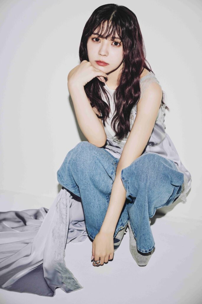 Portrait of a young woman with long dark hair resting her chin on her hand, wearing a white top and blue jeans against a light background.