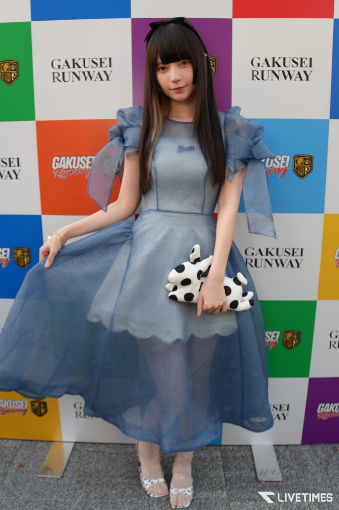 Young woman in a blue puff-sleeve dress posing in front of a colorful backdrop that says 'GAKUSEI RUNWAY', holding a white polka-dot stuffed bag.