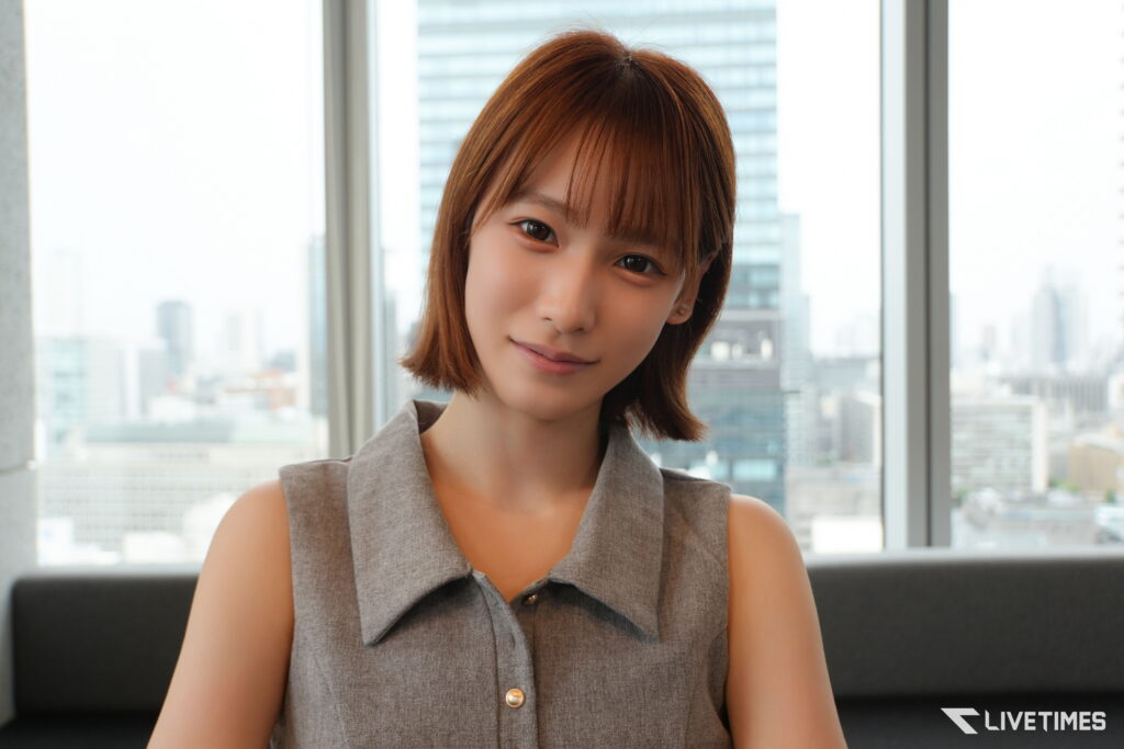 Close-up portrait of a woman with short brown hair, smiling, wearing a gray sleeveless top, with a cityscape visible through large windows in the background.