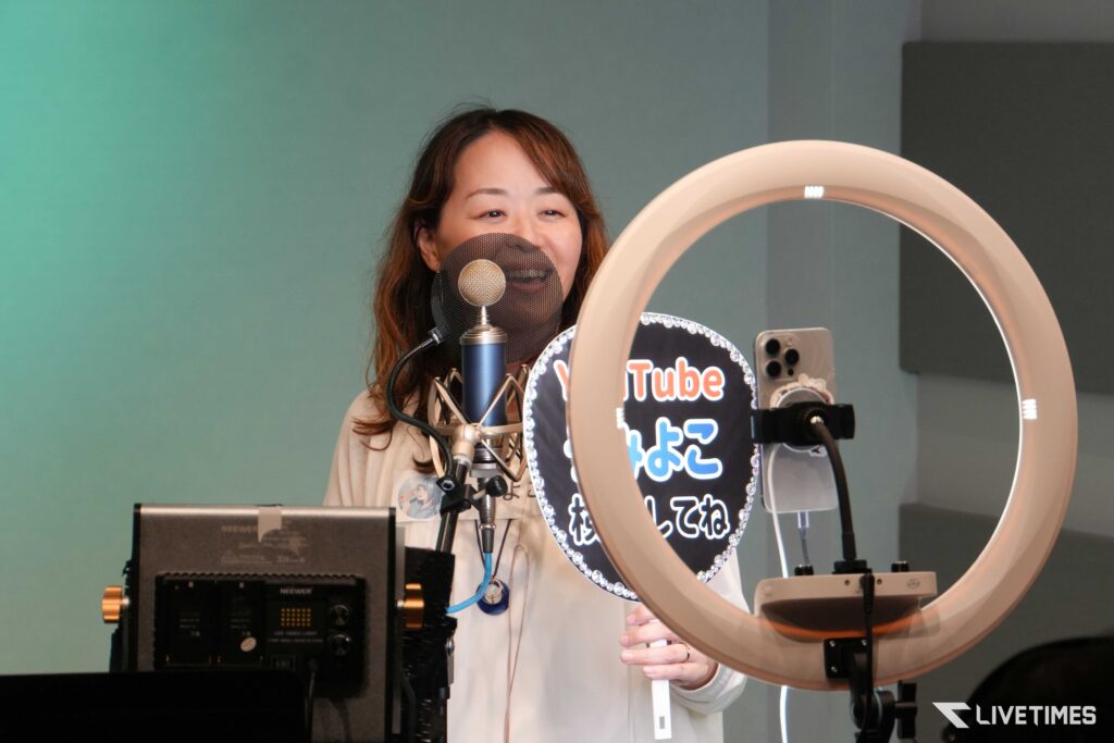 Woman recording vocals in a studio, speaking into a microphone with a pop filter, large circular ring light nearby and a teal wall in the background, Livetimes branding visible.