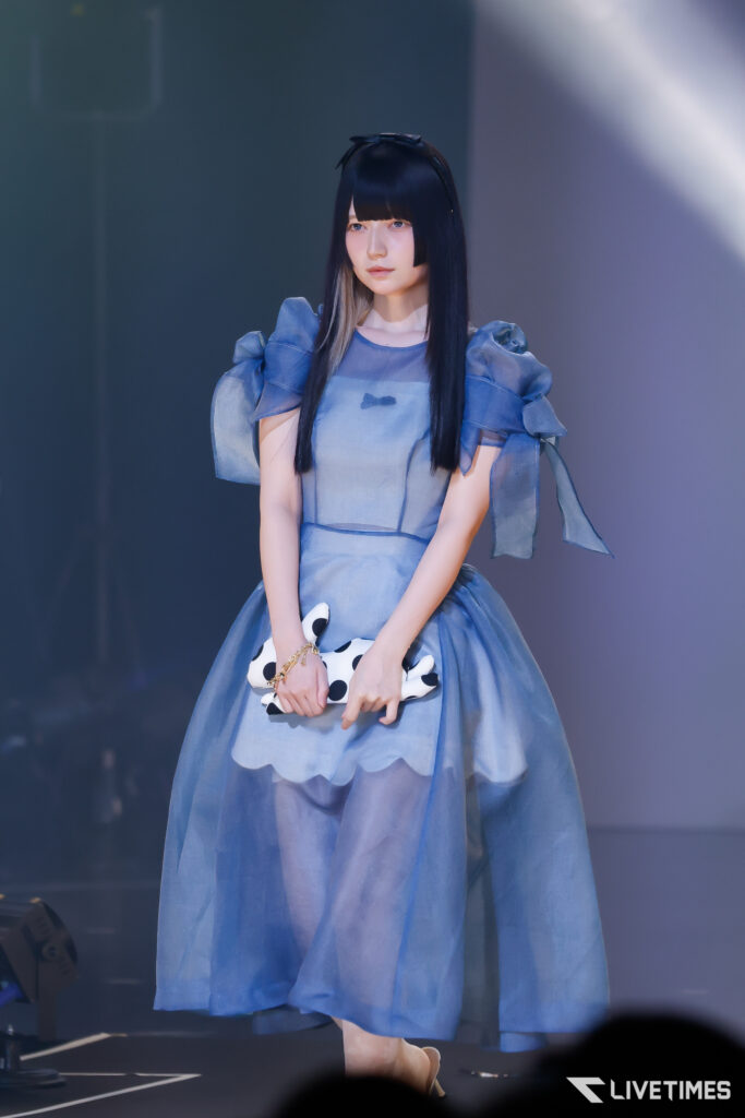 Young woman in a blue puff-sleeve dress standing on a stage, holding a white black-spotted clutch in front of her under blue spotlight
