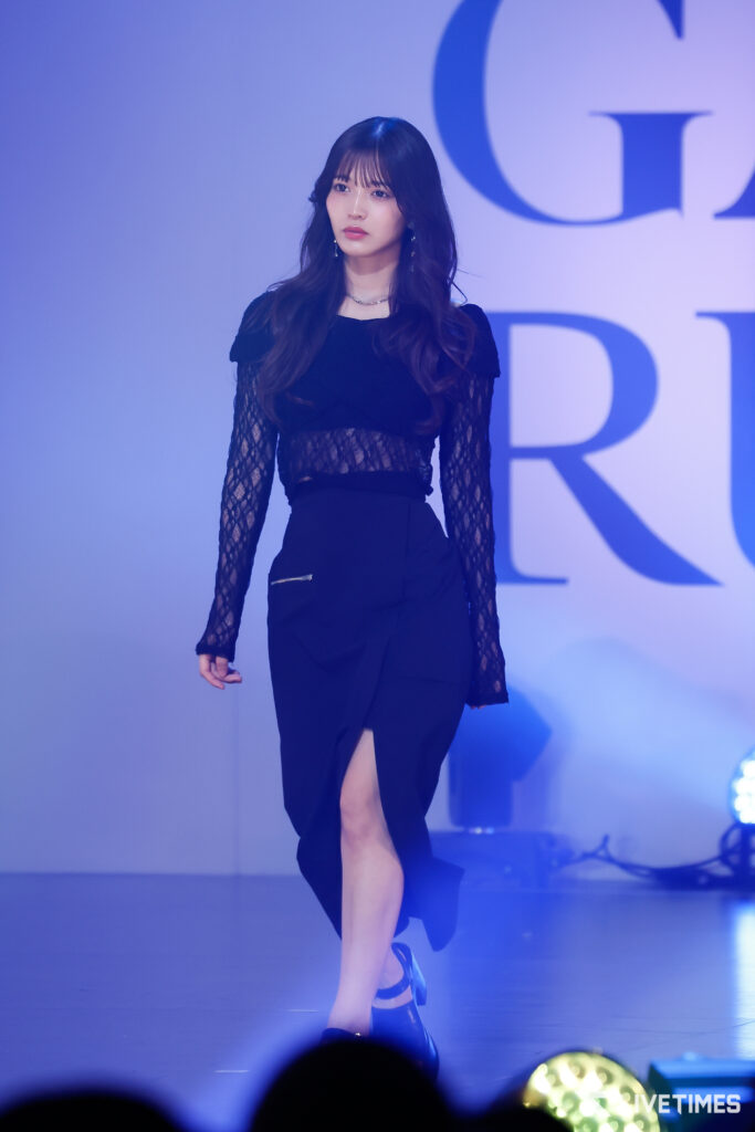 Model walks the runway in a black lace-sleeve dress under blue stage lighting with a focused expression.