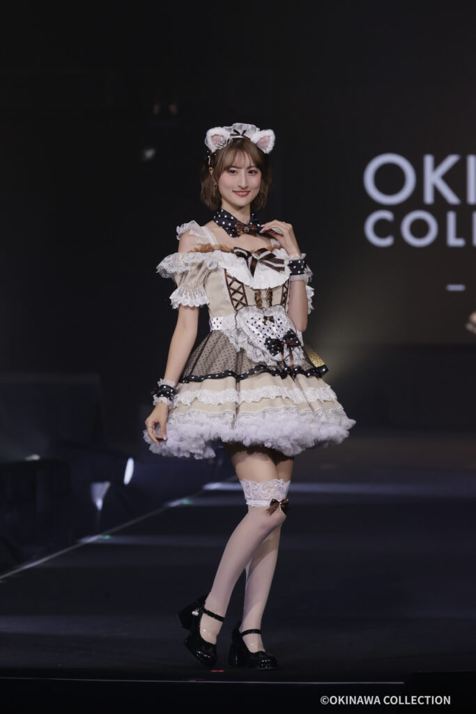 Young woman in a frilly Lolita-style beige and white dress with lace, cat ears, and bows posing on a stage. Text 'OKINAWA COLLECTION' visible in background.