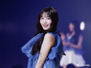 Smiling female performer in a blue dress on a stage with a purple backdrop.