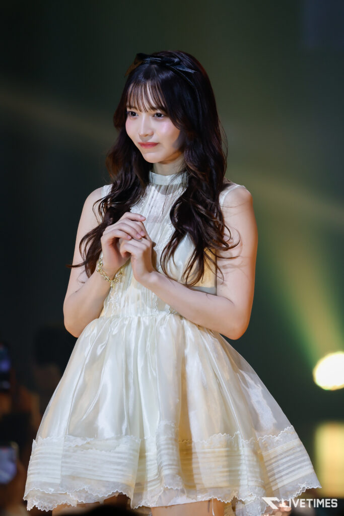 Young woman in a white, lace-trimmed dress on stage, hands clasped, soft spotlight and blurred audience in the background
