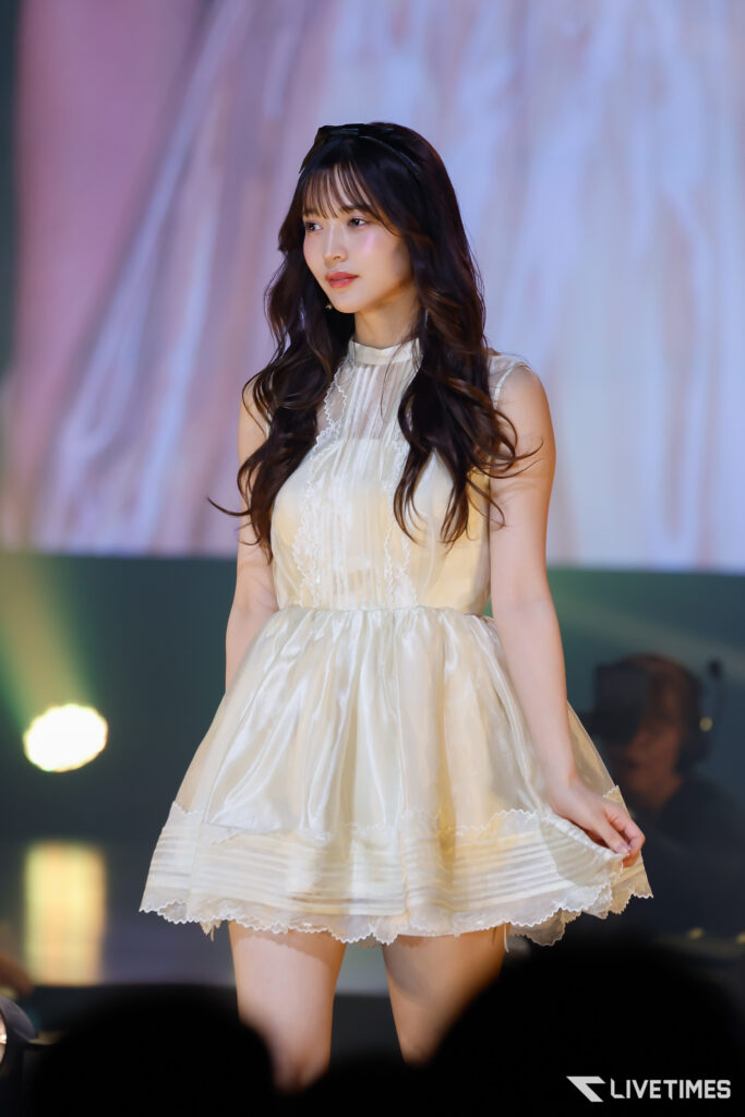 Young woman in a cream dress standing on a stage with soft, colorful lights in the background.
