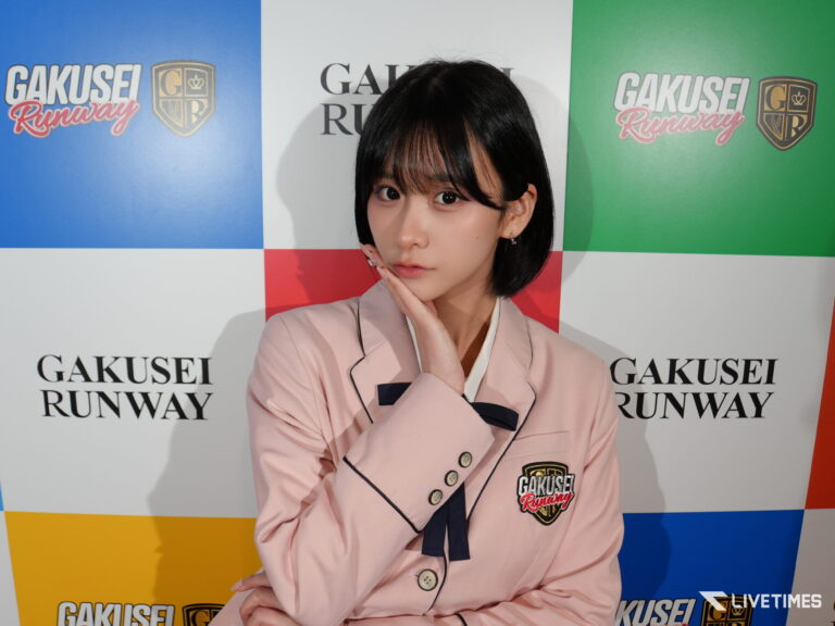 Model posing in a pink blazer with her hand under her chin, in front of a colorful GAKUSEI RUNWAY backdrop with blue, white, red, green, and yellow panels and logos.