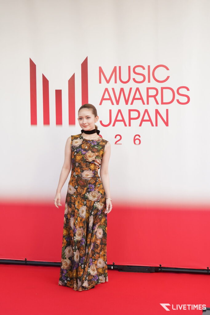 Woman in a long floral dress posing on a red carpet in front of a 'Music Awards Japan 2016' backdrop.