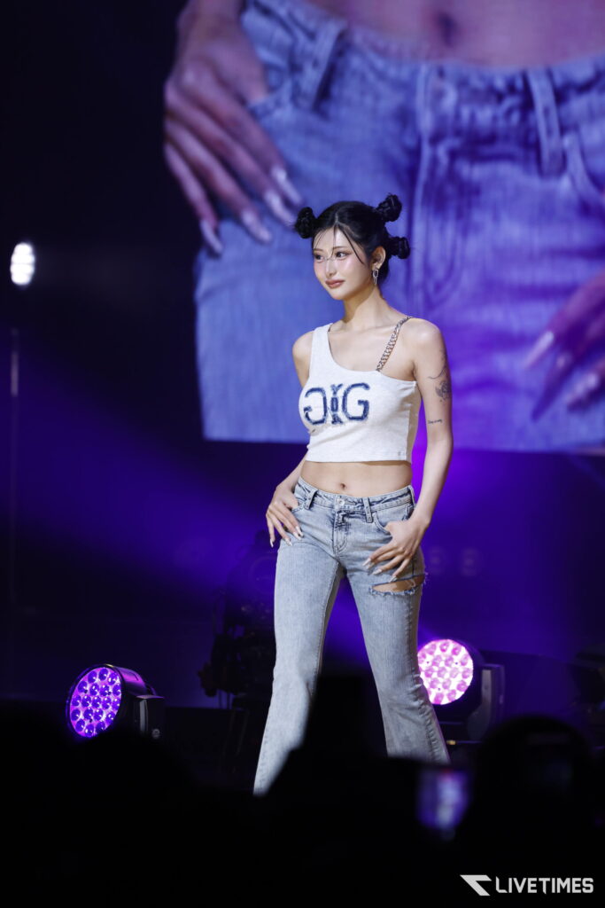 Young woman on stage in a white cropped top and ripped jeans, purple stage lights in the background.