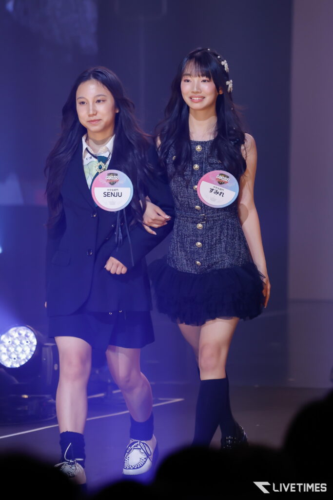 Two young performers walk arm in arm on a lit stage, wearing stage outfits and round badge tags (one reads SENJU).