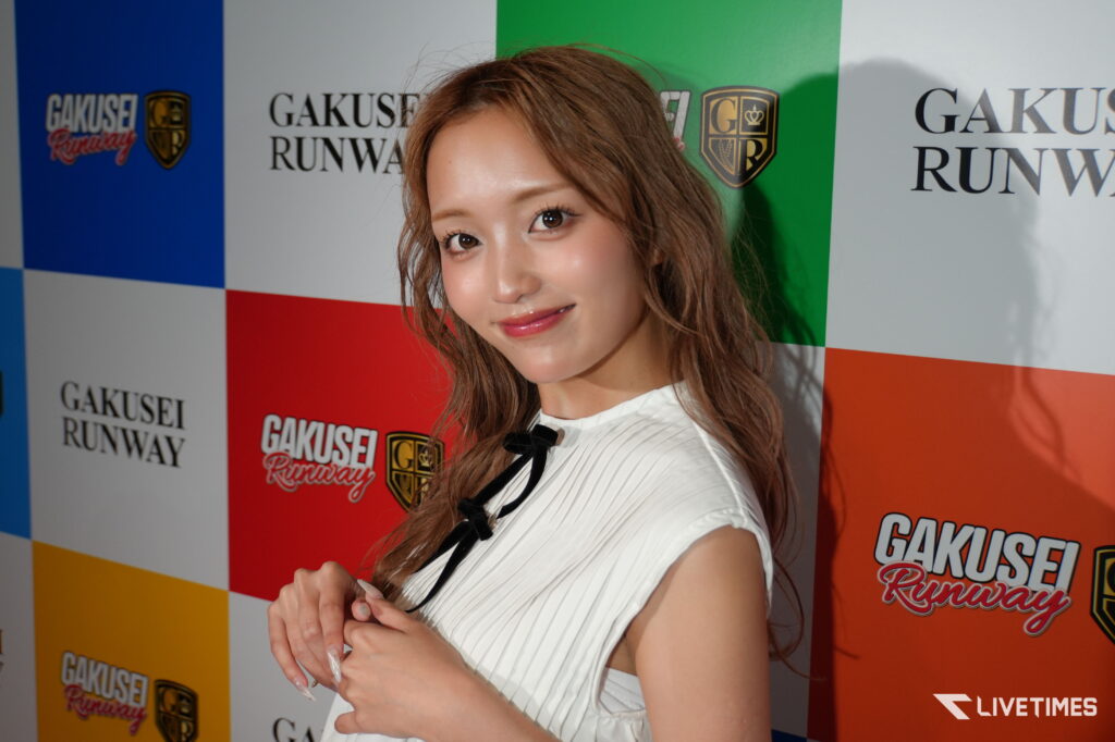 Smiling young woman posing at a Gakusei Runway event in a white striped top against a colorful branded backdrop.