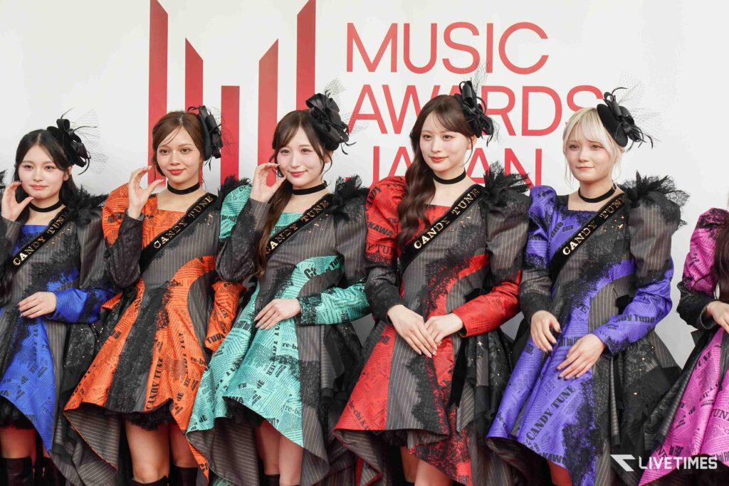 Five women in colorful printed outfits with black sashes posing on a red-carpet backdrop that reads Music Awards Japan.