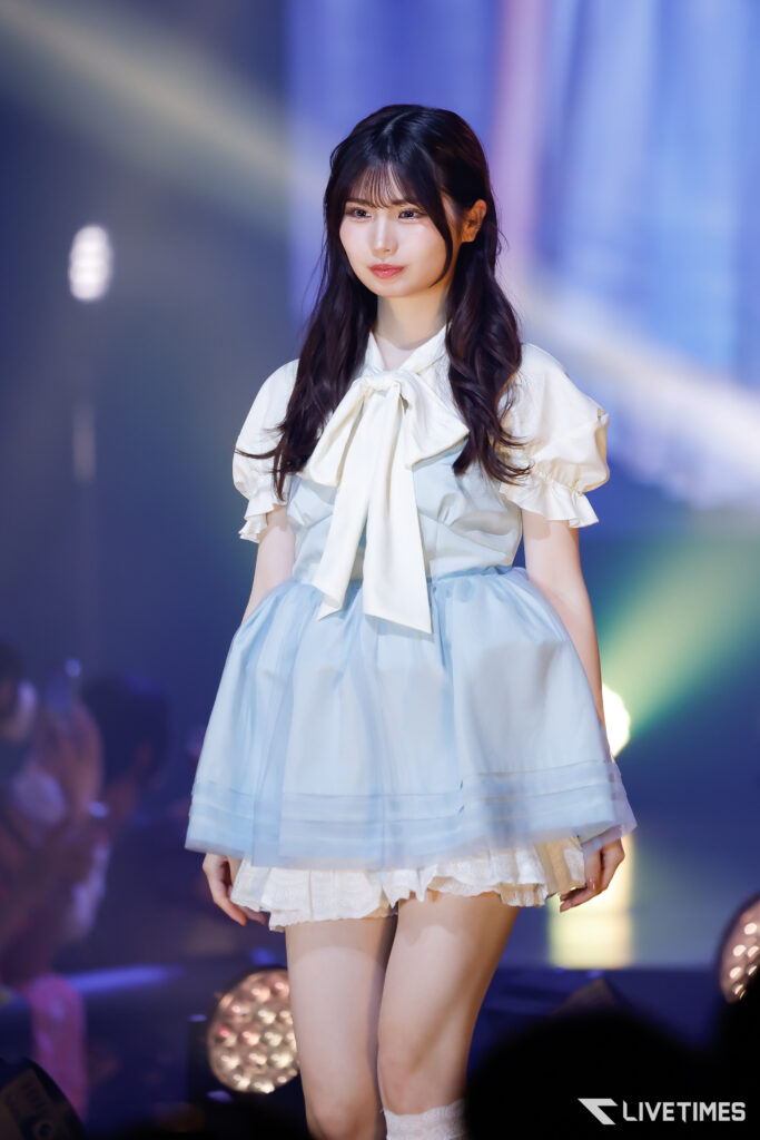 Performer on stage wearing a pale blue dress with a large white bow, under blue stage lights.