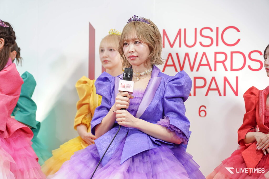 Young woman in a purple gown with a tiara speaks into a microphone at a music awards event