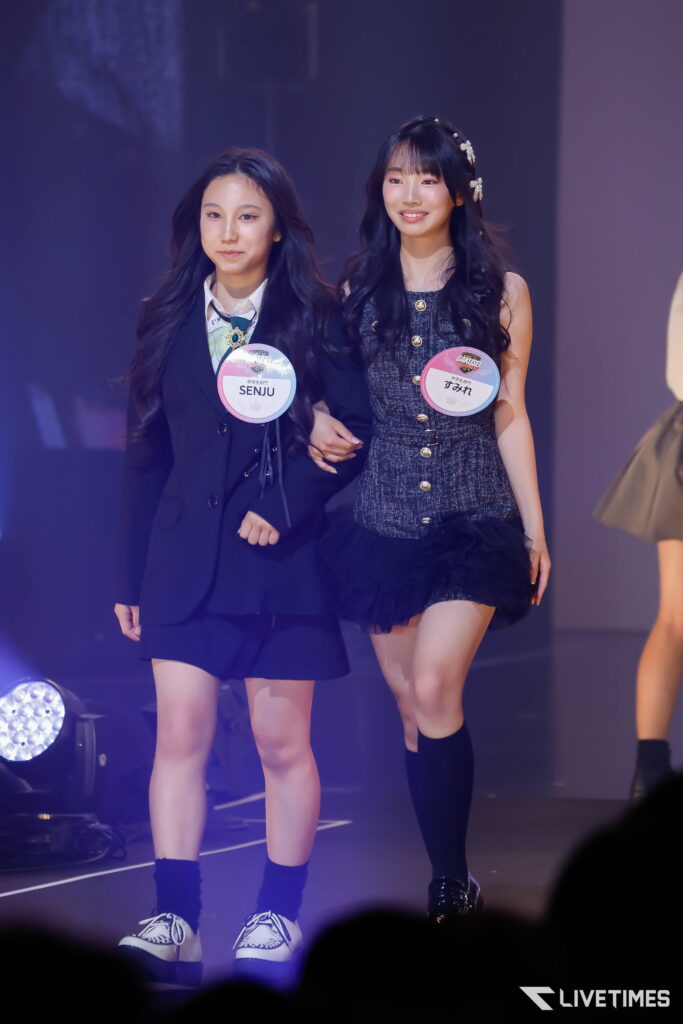 Two young women walk on a stage wearing name badges; one in a dark blazer and skirt, the other in a sparkly dress with hair accessories.