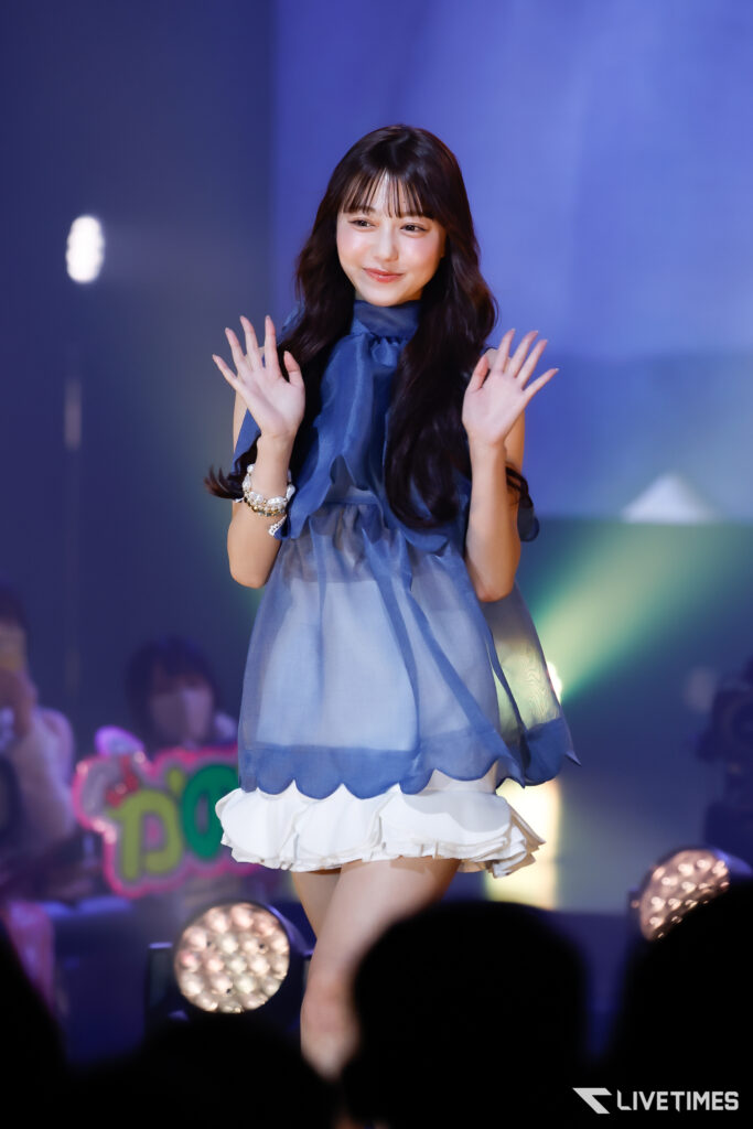 Young woman on stage in a blue sheer top waves to the audience under colorful lights.