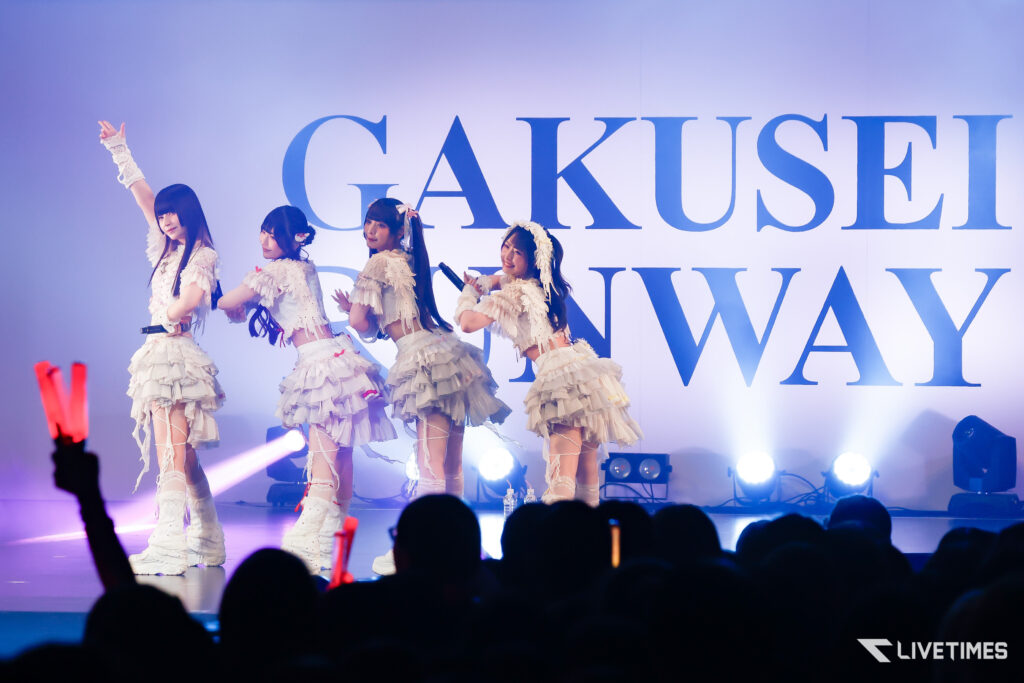 Four female performers in white frilly outfits on stage, posing with synchronized gestures in front of a large 'GAKUSEI' banner and bright stage lights.