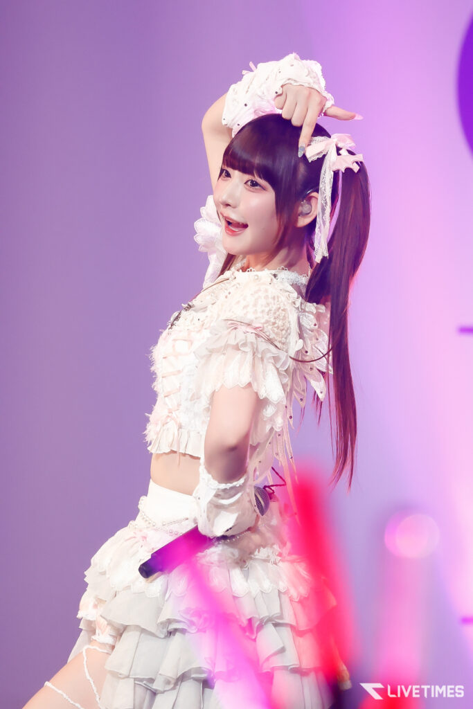 Young female idol performing on stage in a white frilly outfit, arm raised, under pink-purple lights.