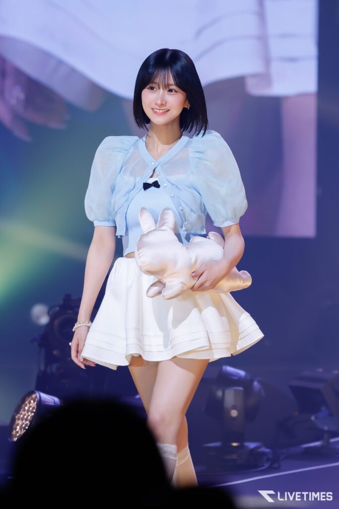 Young female performer on a stage, wearing a light blue puff-sleeve top and white skirt, smiling while holding a large white prop under colorful lights.
