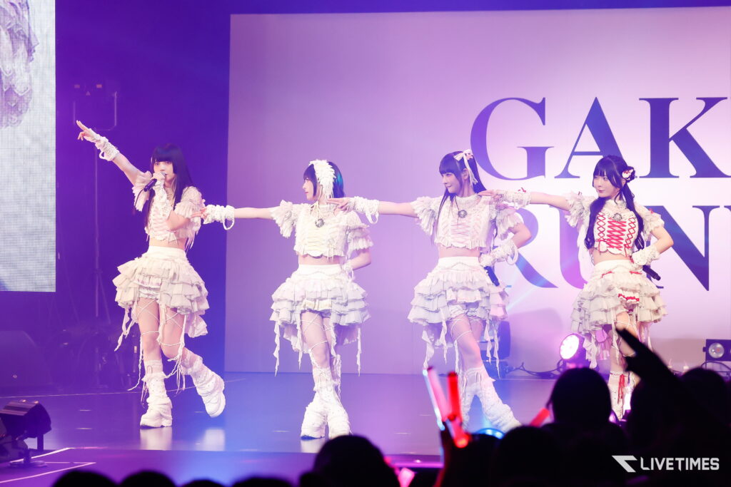 Four female performers in frilly white outfits dance in a line on stage, purple lighting and a large backdrop behind them. Audience with glow sticks visible in the foreground.