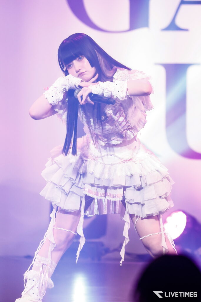 Female performer on stage in a ruffled white-pink dress, holding a microphone and resting her chin on her hand under pink-purple lights