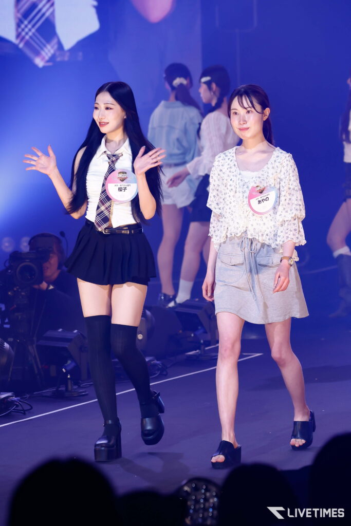 Two young women walk on a dimly lit stage with blue lighting, wearing name badges; one in a white blouse and plaid tie, the other in a floral top and gray skirt, with audience silhouettes in the foreground.