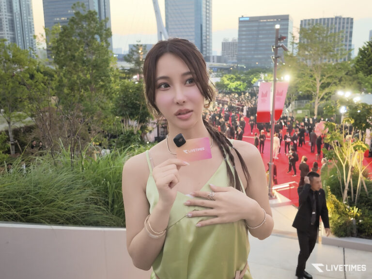 Young woman in a pale green dress posing on a red carpet outdoors, holding a pink LIVETIME card with a headset mic clipped to her chest, city skyline in the background.