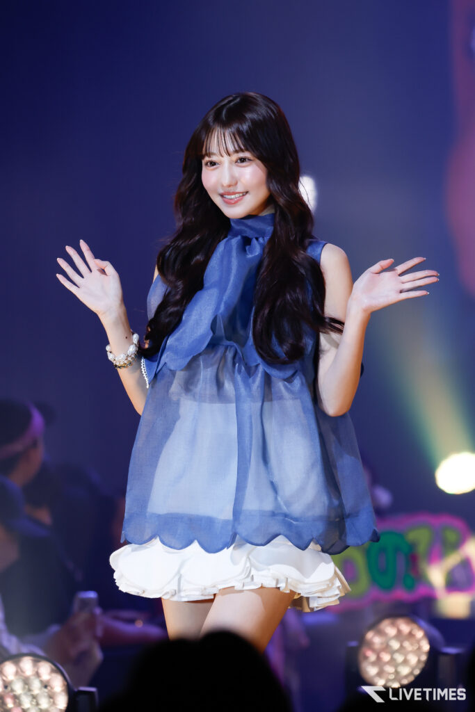 Young woman on stage waving both hands, wearing a blue sleeveless top with a scarf and white ruffled skirt under stage lights.